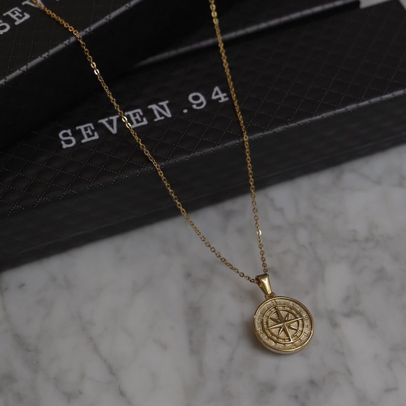 May include: A gold compass necklace with a delicate chain. The compass pendant features a detailed design with a star in the center and the points of the compass marked around the edge. The necklace is displayed on a black box with the text "SEVEN.94" printed on it.