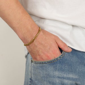 Peut inclure: Un bracelet en cha&icirc;ne dor&eacute;e au poignet d'une personne. Le bracelet est fait d'une cha&icirc;ne &eacute;paisse de style curb.