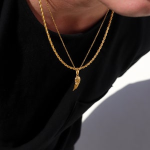 Puede incluir: Un collar de cadena dorada con un colgante de ala de &aacute;ngel dorada. El collar est&aacute; superpuesto a una camiseta negra.