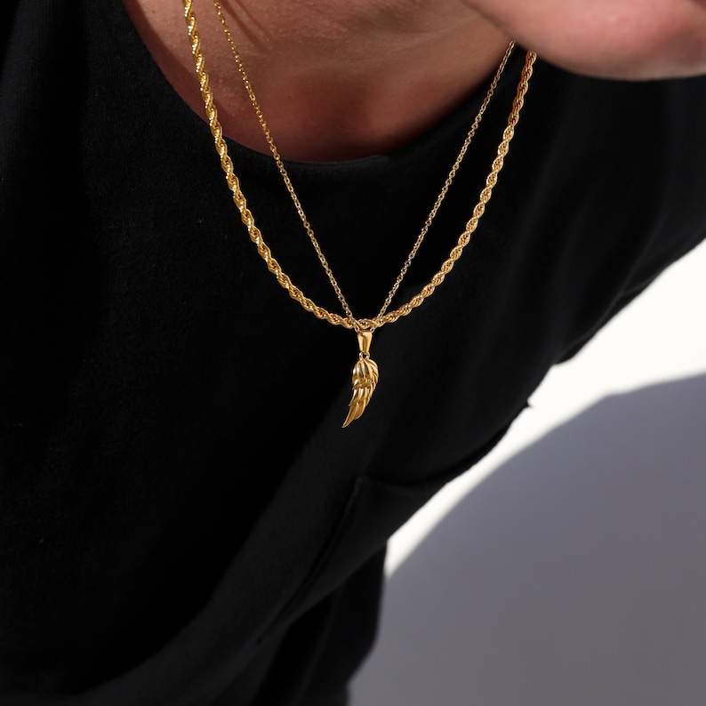 Puede incluir: Un collar de cadena dorada con un colgante de ala de &aacute;ngel dorada. El collar est&aacute; superpuesto a una camiseta negra.