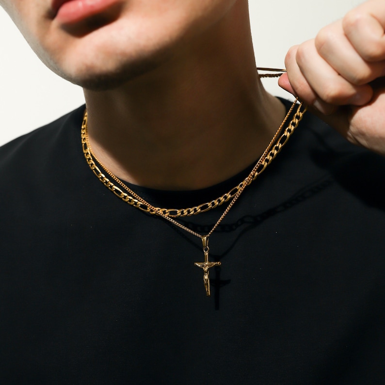 May include: A gold chain necklace with a gold cross pendant. The chain is a layered design with a thicker, more prominent chain on top of a thinner chain.