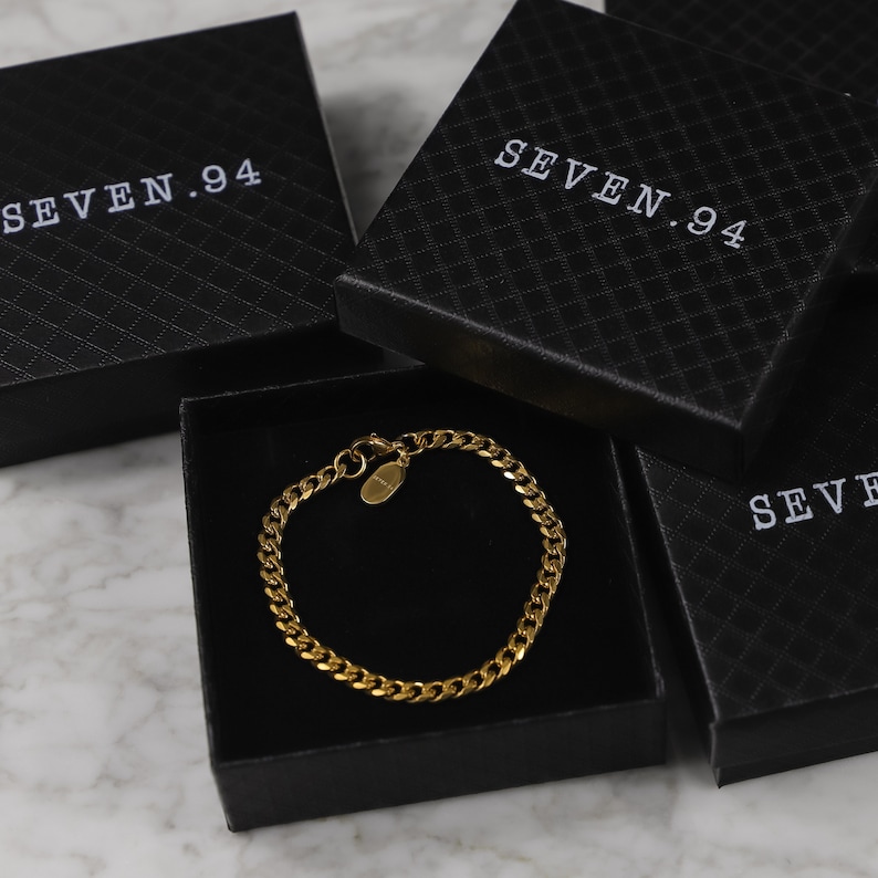 Peut inclure: Un bracelet en cha&icirc;ne dor&eacute;e avec une petite &eacute;tiquette ovale, pr&eacute;sent&eacute; dans une bo&icirc;te noire avec le texte "SEVEN.94" imprim&eacute; sur le couvercle.