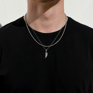 Puede incluir: Un collar de cadena de plata con un colgante de ala de plata. El collar est&aacute; superpuesto a una cadena de plata m&aacute;s fina.
