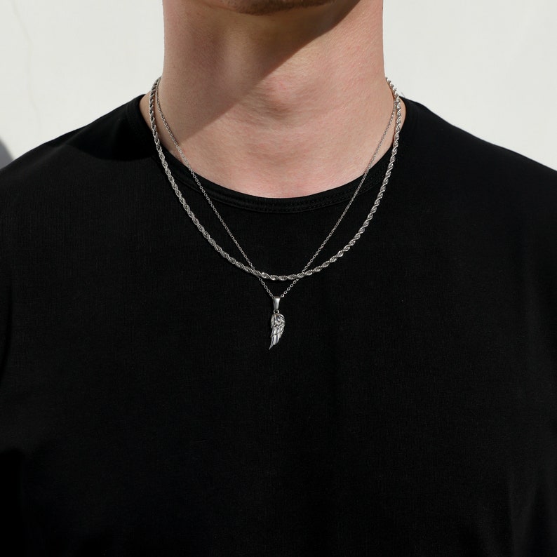 Puede incluir: Un collar de cadena de plata con un colgante de ala de plata. El collar est&aacute; superpuesto a una cadena de plata m&aacute;s fina.