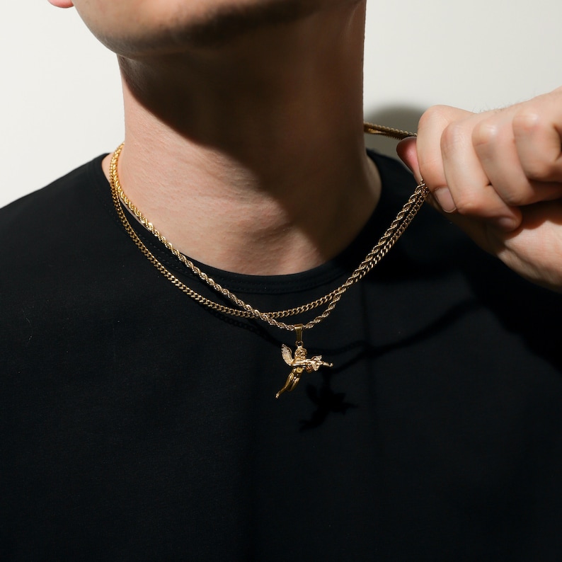 Puede incluir: Un collar de cadena de oro con un peque&ntilde;o colgante de &aacute;ngel. El collar est&aacute; superpuesto con una segunda cadena m&aacute;s gruesa.