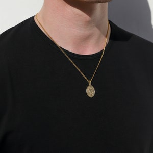 May include: A gold chain necklace with a small oval pendant featuring a detailed design. The pendant is hanging on a person's chest.