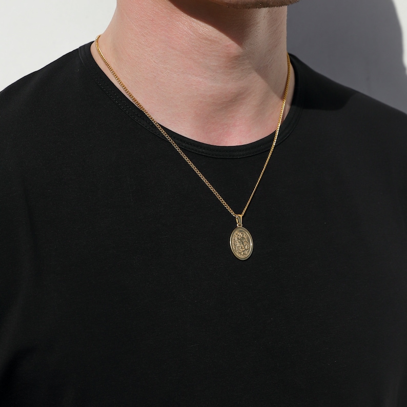 May include: A gold chain necklace with a small oval pendant featuring a detailed design. The pendant is hanging on a person's chest.