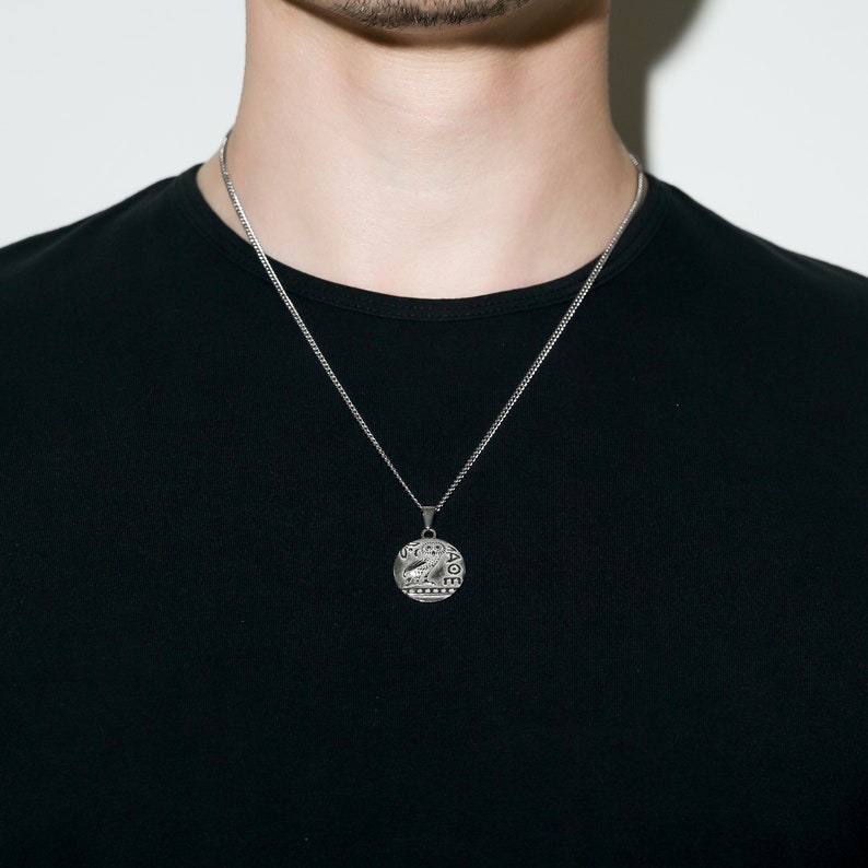 May include: A silver chain necklace with a round pendant featuring a detailed design. The pendant has a raised image of an owl with text around the edge.