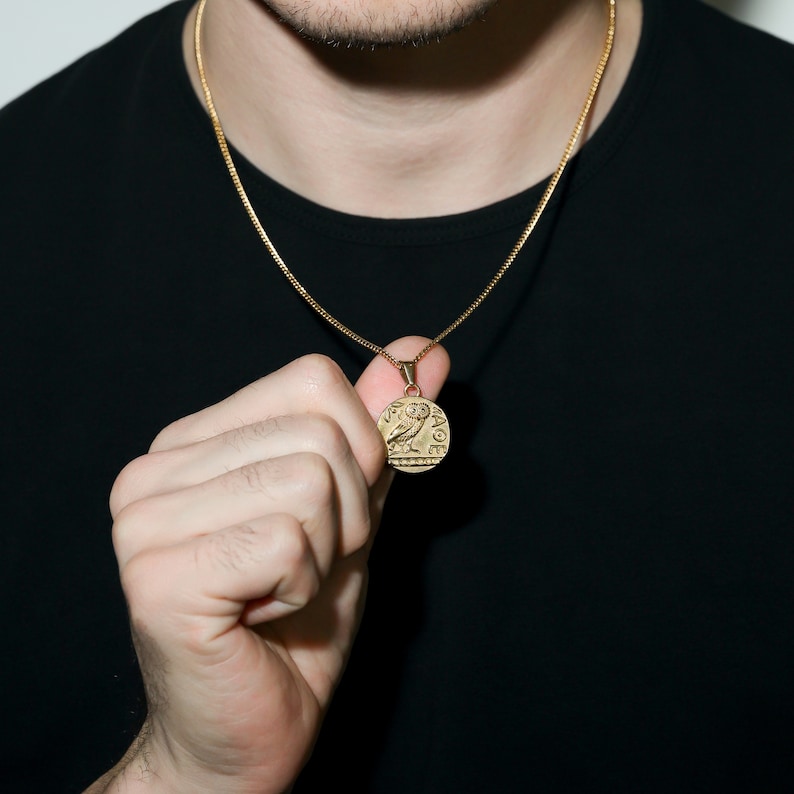 May include: A gold chain necklace with a round pendant featuring an owl design. The pendant has intricate details and appears to be an ancient Greek coin.