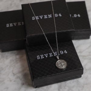 May include: A silver compass necklace with a round pendant on a black box with the text "SEVEN.94" printed on it.