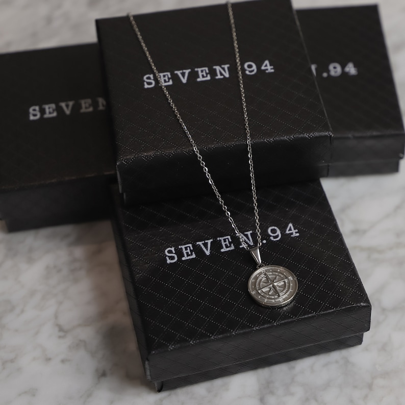 May include: A silver compass necklace with a round pendant on a black box with the text "SEVEN.94" printed on it.