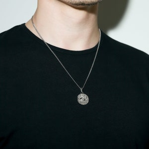 May include: A silver chain necklace with a round pendant featuring a detailed design. The pendant has the word "GRACE" inscribed around the edge.