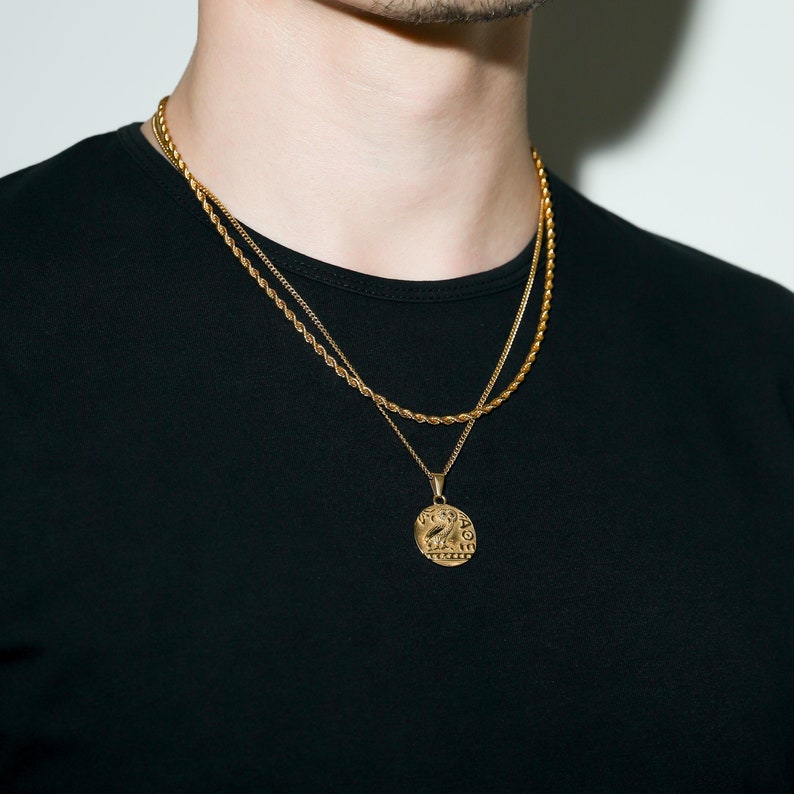 May include: A gold chain necklace with a gold coin pendant featuring a detailed design. The necklace is layered over a second, thinner gold chain.