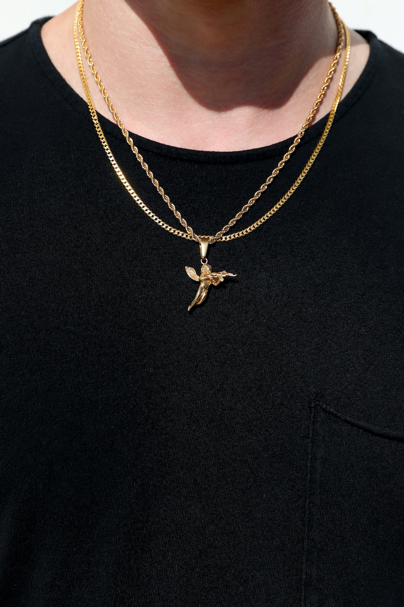 Puede incluir: Un collar de cadena de oro con un peque&ntilde;o colgante de &aacute;ngel. El collar est&aacute; superpuesto con una segunda cadena m&aacute;s gruesa.