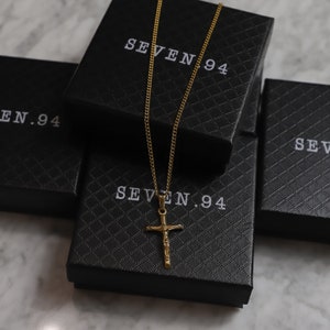 May include: A gold chain necklace with a gold cross pendant. The necklace is displayed on top of three black boxes with the text "SEVEN.94" printed on them.