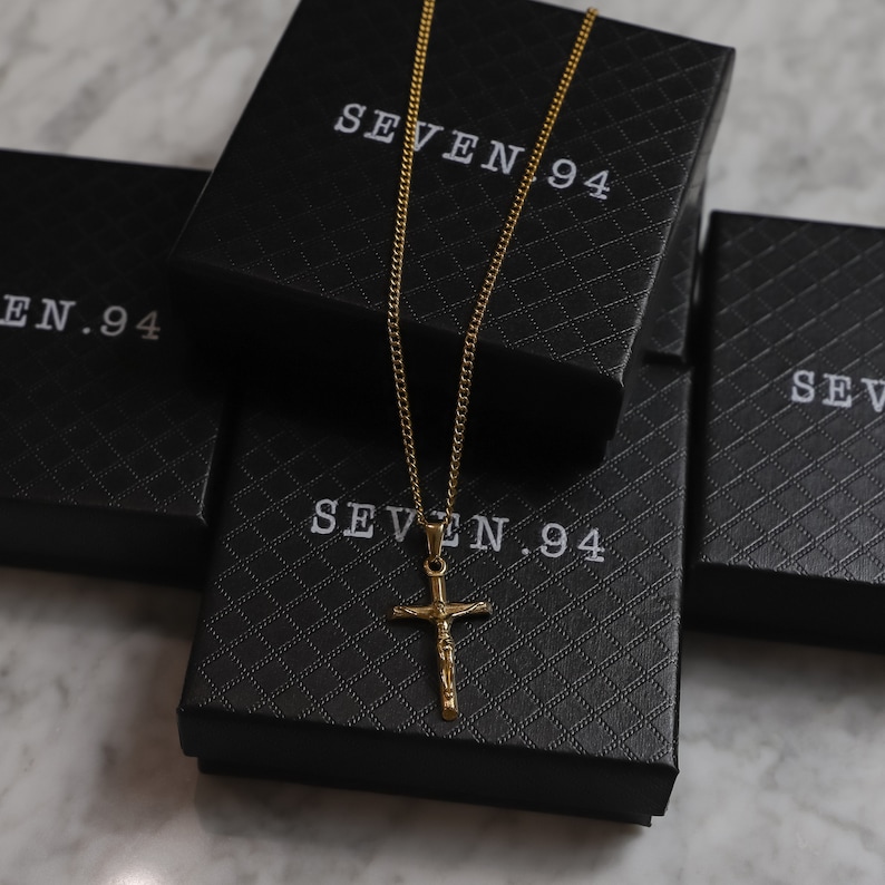 May include: A gold chain necklace with a gold cross pendant. The necklace is displayed on top of three black boxes with the text "SEVEN.94" printed on them.