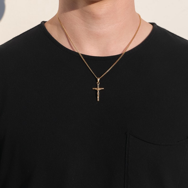 May include: A gold chain necklace with a small gold cross pendant. The cross features a detailed design of a figure on it.