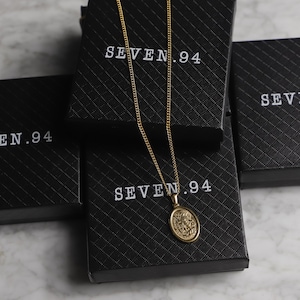 May include: A gold chain necklace with an oval pendant featuring a detailed design. The necklace is displayed on a black box with the text "SEVEN.94" printed on it.