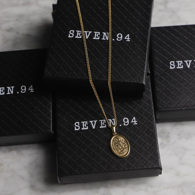 May include: A gold chain necklace with an oval pendant featuring a detailed design. The necklace is displayed on a black box with the text "SEVEN.94" printed on it.