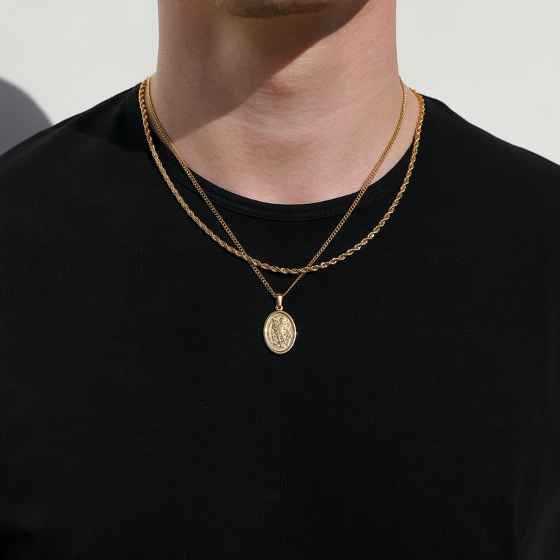May include: A gold chain necklace with a rope chain and a pendant featuring a religious figure. The necklace is layered over a second, thinner chain.
