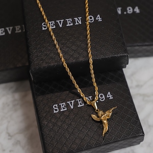 Puede incluir: Un collar de cadena de oro con un colgante de &aacute;ngel de oro que sostiene una pistola. El collar se muestra sobre una caja negra con el texto "SEVEN 94" impreso en ella.