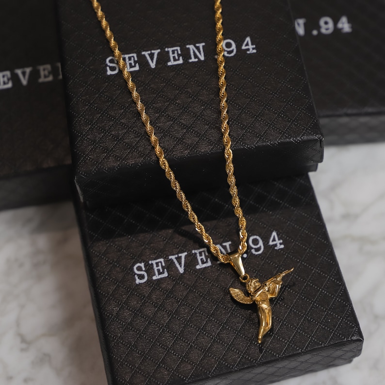 Puede incluir: Un collar de cadena de oro con un colgante de &aacute;ngel de oro que sostiene una pistola. El collar se muestra sobre una caja negra con el texto "SEVEN 94" impreso en ella.