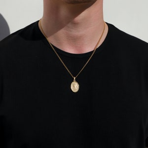 May include: A gold chain necklace with a small oval pendant featuring a detailed design. The necklace is worn by a person with a black shirt.