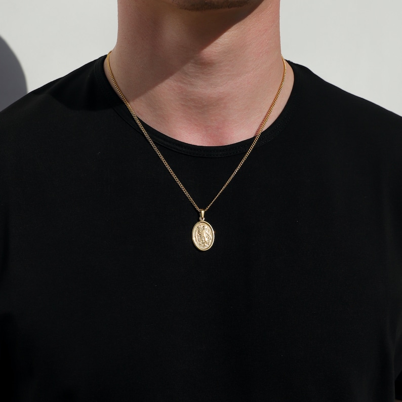 May include: A gold chain necklace with a small oval pendant featuring a detailed design. The necklace is worn by a person with a black shirt.