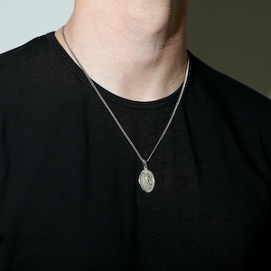 May include: A silver chain necklace with an oval pendant featuring a detailed design. The necklace is worn by a person wearing a black t-shirt.