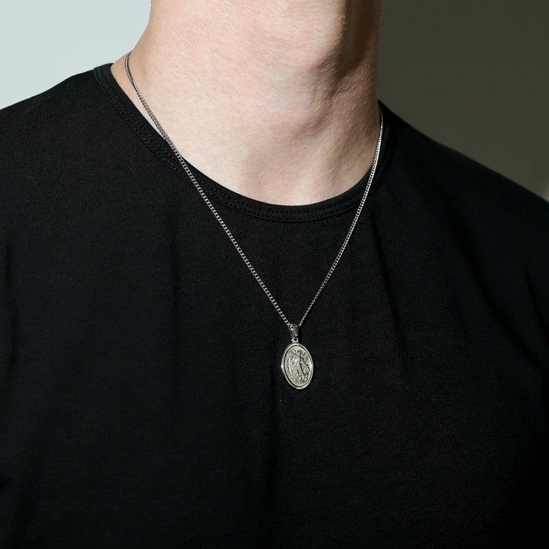 May include: A silver chain necklace with an oval pendant featuring a detailed design. The necklace is worn by a person wearing a black t-shirt.