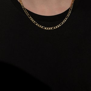 May include: A gold chain necklace with a classic figaro design. The chain is worn by a person with a black shirt.