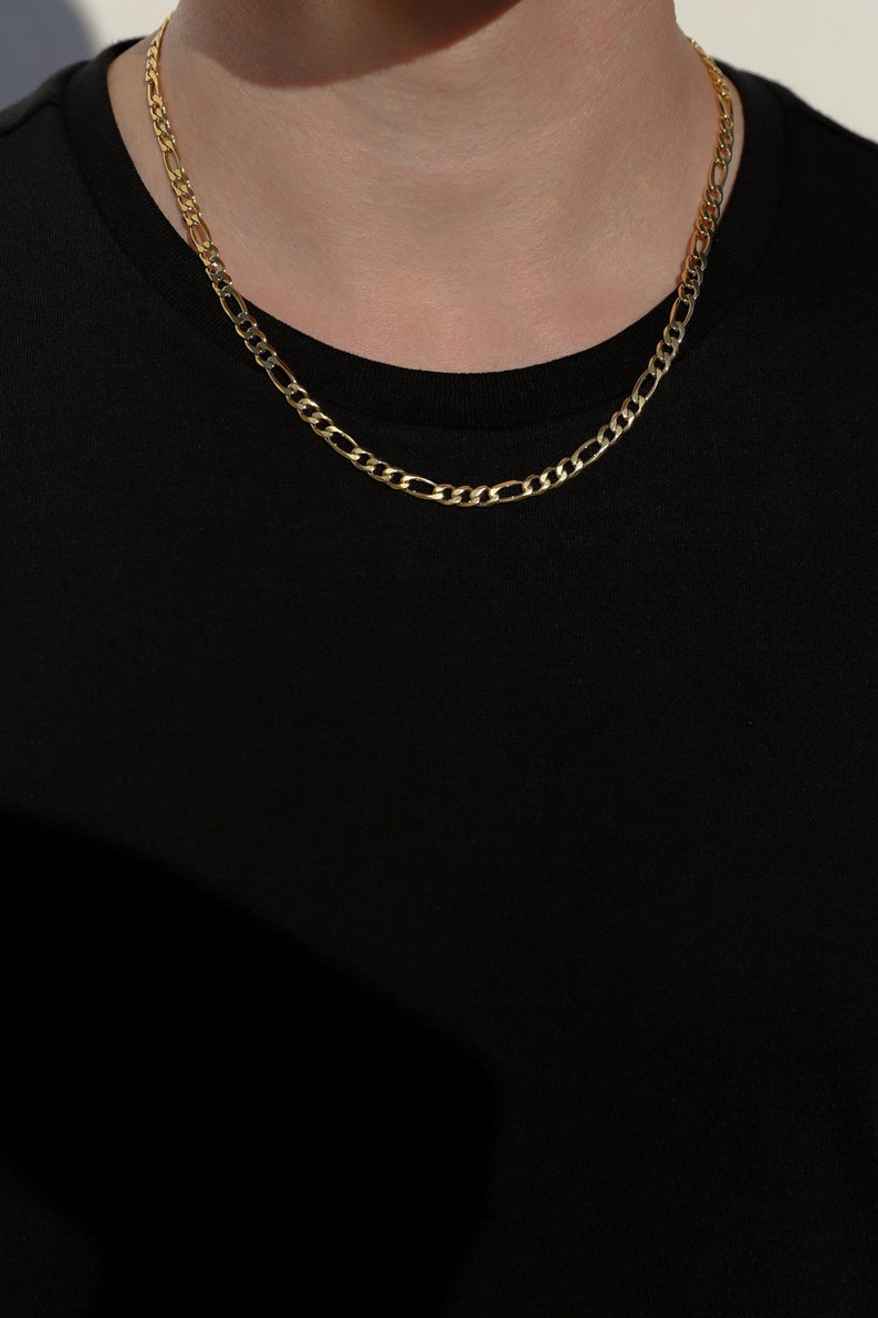 May include: A gold chain necklace with a classic figaro design. The chain is worn by a person with a black shirt.