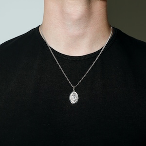 May include: A silver chain necklace with a silver oval pendant featuring a detailed design.