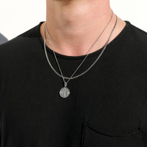 May include: A silver chain necklace with a silver pendant featuring a circular design. The necklace is layered on top of another silver chain.