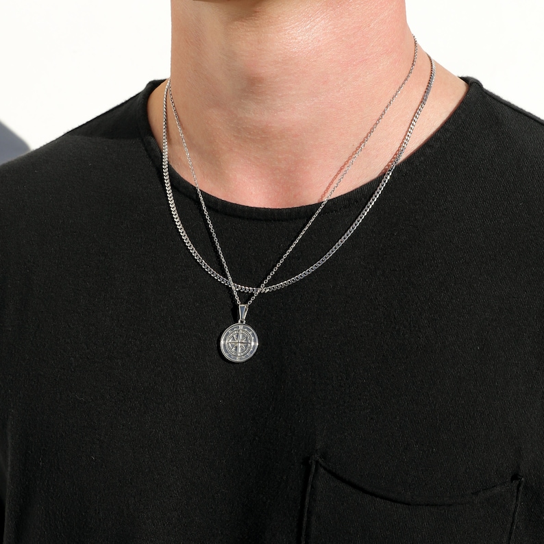 May include: A silver chain necklace with a silver pendant featuring a circular design. The necklace is layered on top of another silver chain.