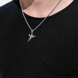 Puede incluir: Un collar de cadena de plata con un colgante de querub&iacute;n de plata. El collar lo lleva una persona que lleva una camisa negra.