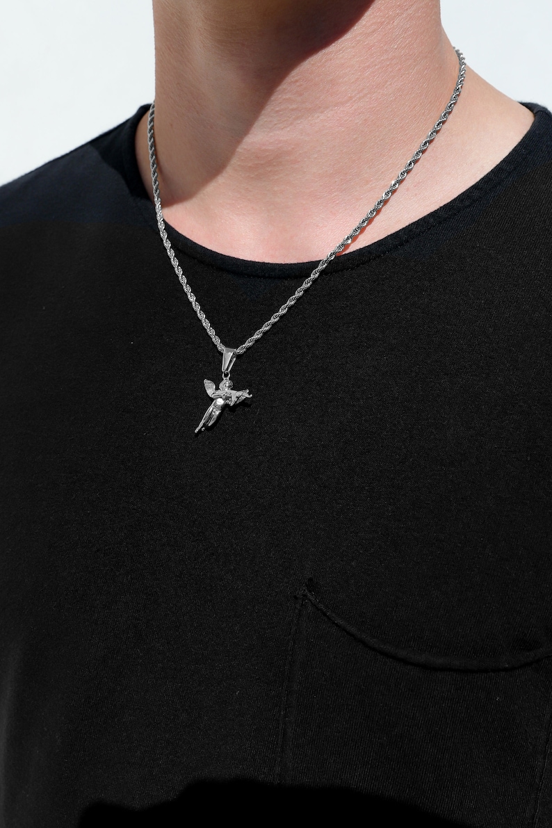 Puede incluir: Un collar de cadena de plata con un colgante de querub&iacute;n de plata. El collar lo lleva una persona que lleva una camisa negra.
