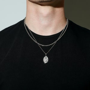 May include: A silver chain necklace with a silver pendant featuring a detailed design. The necklace is layered over a second, thinner silver chain.