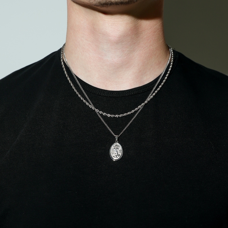 May include: A silver chain necklace with a silver pendant featuring a detailed design. The necklace is layered over a second, thinner silver chain.