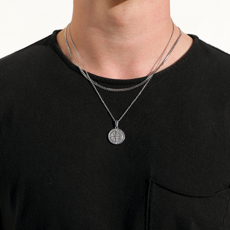 May include: A silver chain necklace with a round pendant featuring a compass design. The necklace is layered with two other silver chains.