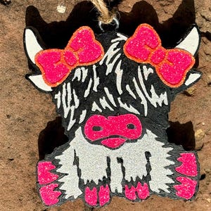 Puede incluir: Adorno de madera de una vaca de las Tierras Altas con una cuerda de yute para colgar. La vaca tiene un cuerpo negro, cara y patas blancas, y detalles en rosa brillante en los lazos, la nariz y las pezu&ntilde;as. El adorno est&aacute; sobre un fondo marr&oacute;n texturizado.