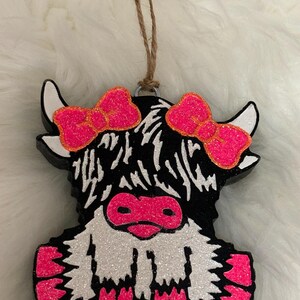 Puede incluir: Adorno decorativo con forma de vaca de las Tierras Altas, con un cuerpo negro y blanco y detalles de purpurina rosa. La vaca tiene lazos rosas y una nariz rosa. Se adjunta una cuerda de yute para colgar.
