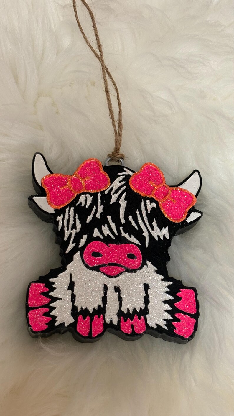 Puede incluir: Adorno decorativo con forma de vaca de las Tierras Altas, con un cuerpo negro y blanco y detalles de purpurina rosa. La vaca tiene lazos rosas y una nariz rosa. Se adjunta una cuerda de yute para colgar.