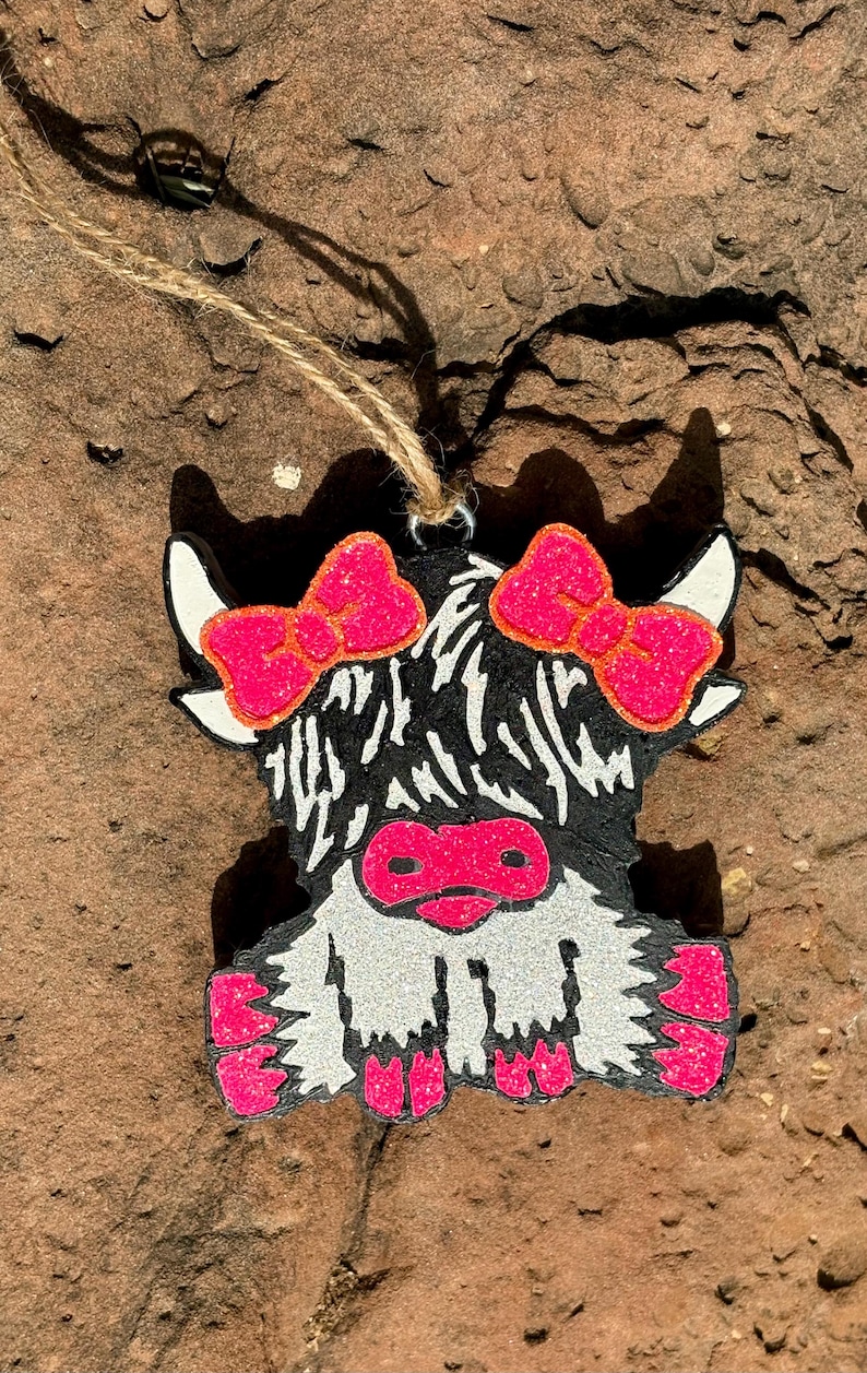 Puede incluir: Adorno de madera de una vaca de las Tierras Altas con una cuerda de yute para colgar. La vaca tiene un cuerpo negro, cara y patas blancas, y detalles en rosa brillante en los lazos, la nariz y las pezu&ntilde;as. El adorno est&aacute; sobre un fondo marr&oacute;n texturizado.
