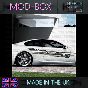 May include: A white car with a black and grey graphic decal on the side. The decal features geometric shapes and lines. The car is parked in front of a building with a sign that says "MOD-BOX".
