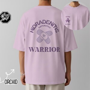May include: A light purple t-shirt featuring a graphic on the back with two crossed adhesive bandages, a heart, and the text "HIDRADENITIS SUPPURATIVA WARRIOR". The front chest area includes a small purple awareness ribbon and the text "TEAM".