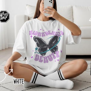May include: A white short-sleeve t-shirt featuring a graphic of a black butterfly encircled by a teal ring, with the purple text "HIDRADENITIS BEAUTY" arched above and below the butterfly, accented by small purple stars and moons.