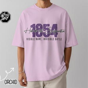 May include: A light purple t-shirt with the text "1854 Hidradenitis Suppurativa VISIBLE NAME. INVISIBLE BATTLE" in dark purple. The shirt has short sleeves and a relaxed fit.