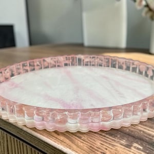 May include: A round, shallow tray with a scalloped edge, featuring a translucent resin construction with a marbled pattern of soft pink and white swirls. The surface is smooth and glossy, with the pink colour concentrated along the ridged rim.