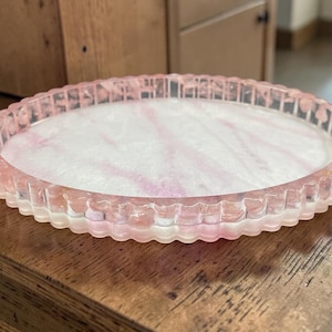 May include: A round, shallow tray with a scalloped edge, crafted from translucent resin. The base features a marbled pattern of soft pink and white swirls, while the raised, fluted rim is a clear, pale pink, creating a cohesive, layered appearance.